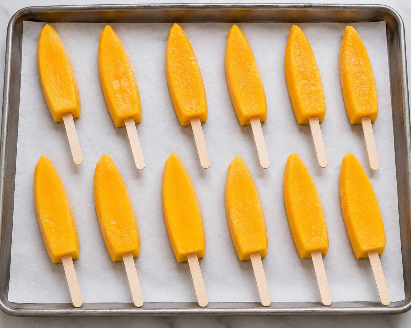 Mango spears on sticks arranged in a single layer on a parchment-lined tray